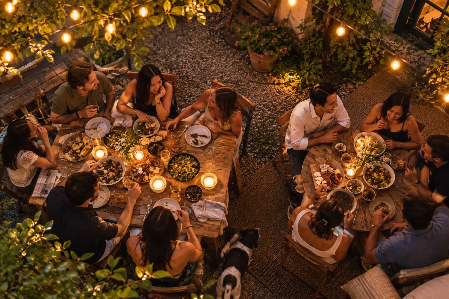 People sharing food and laughing at two candlelit tables in a backyard under string lights.